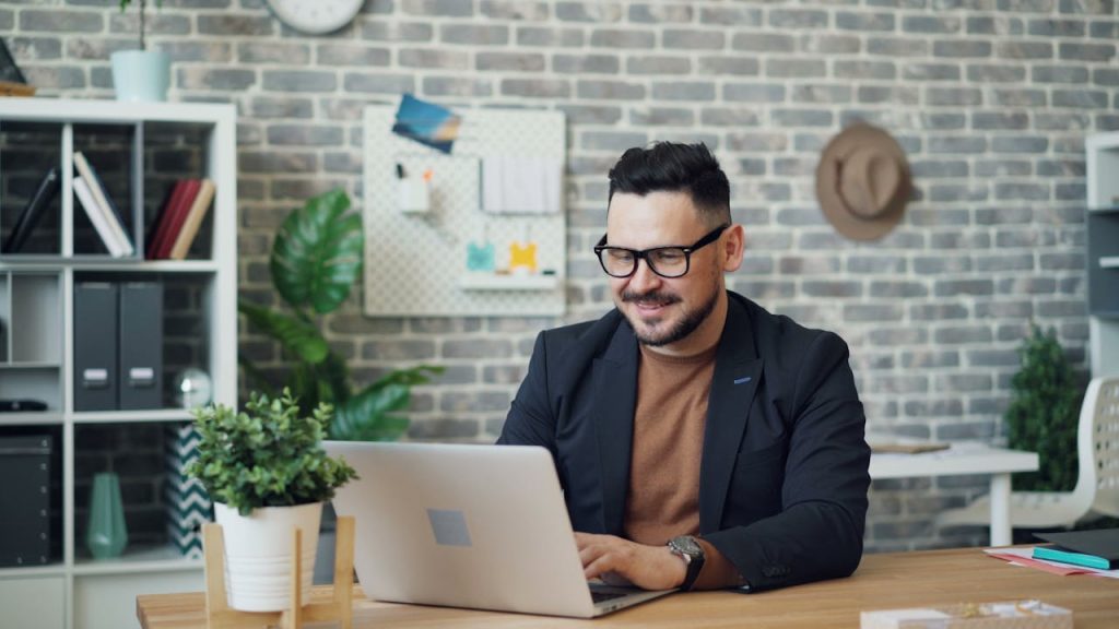 A man smiles while working on a laptop in a stylish, modern office setting.